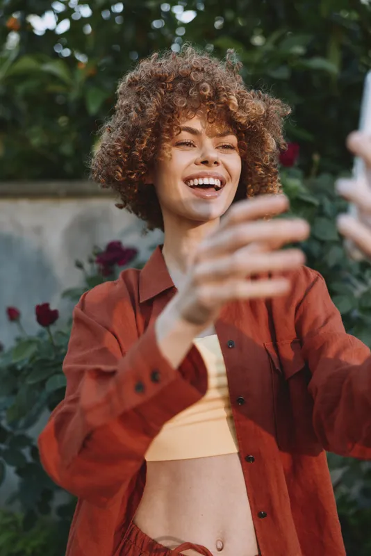 Young woman with curly hair laughs and reaches toward the camera in a sunlit garden, wearing a rust jacket and crop top, conveying carefree summer vibes