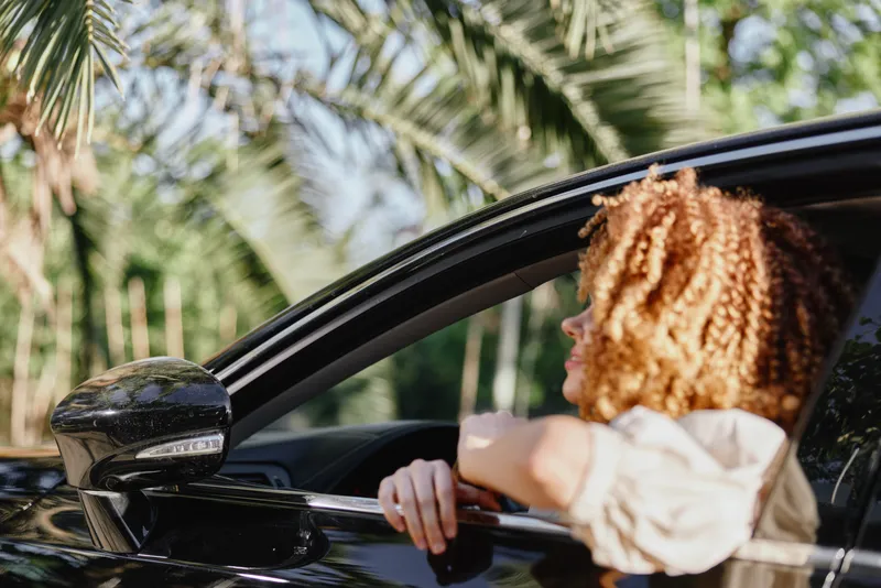 Woman with curly hair looking out of car window, surrounded by tropical palm trees, natural sunlight, and a relaxed summer atmosphere.