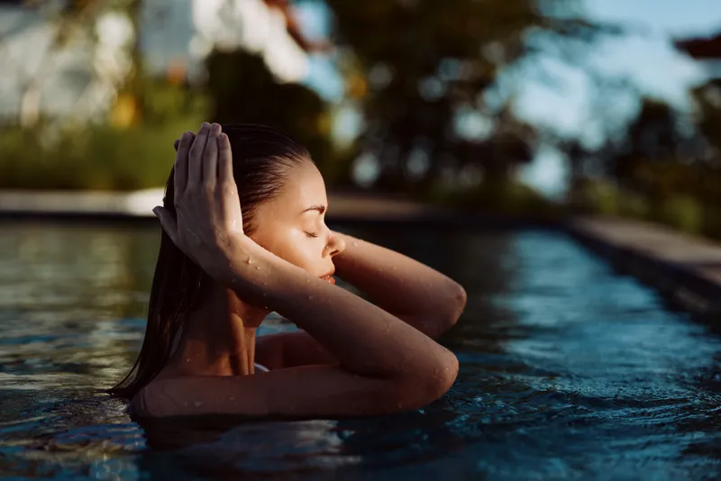 A young woman relaxes in an outdoor pool during sunset with eyes closed and hands behind her head. The calm water and blurred background create a peaceful lifestyle scene.