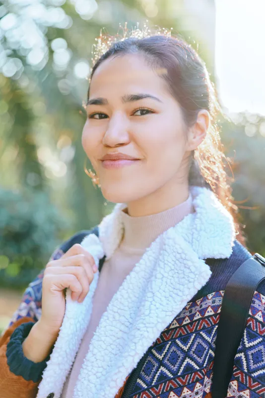 Smiling woman outdoors with backpack, casual fashion and traveler vibe in daylight, standing in a park with warm sunlight, conveying friendly confidence, vibrant color, and everyday lifestyle in