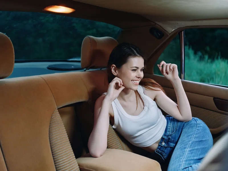 Happy young woman in casual outfit smiling inside vintage car, reflecting joy and freedom, with a serene outdoor background, highlighting relaxation and adventure.