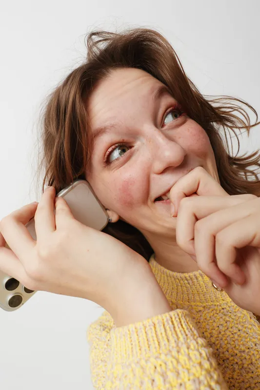 Happy young woman with messy hair holds smartphone near ear smiling and looking away. Casual lifestyle, communication, technology, and positive emotions concept in studio with white background.