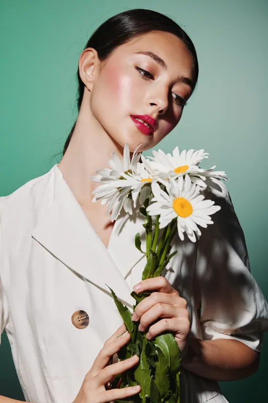 Young woman with glossy skin and sleek hair holds white daisies close, wearing a minimalist beige blazer. Studio portrait shot on a vivid green background about beauty, fashion, and creativity.
