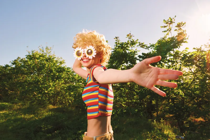A playful child in a striped top wears flower sunglasses and reaches toward the camera in a sunlit field, radiating joyful energy and a retro outdoor vibe, carefree and bright.
