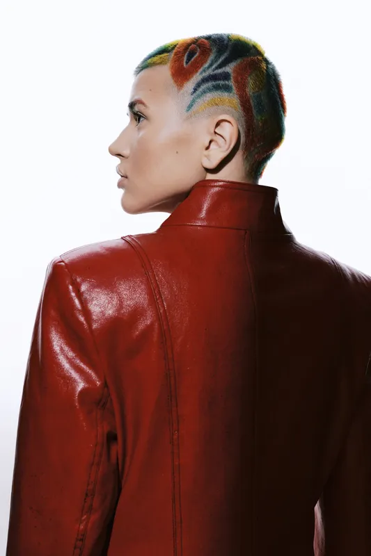 Woman with colorful shaved hair wearing a striking red leather jacket, showcasing unique fashion and bold hairstyle against a white background.