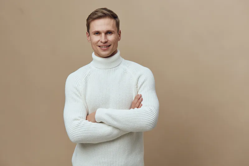 Smiling man wearing white knitted turtleneck sweater posing confidently against beige background. Studio portrait of cheerful young adult male with arms crossed, casual style, lifestyle concept, indoo