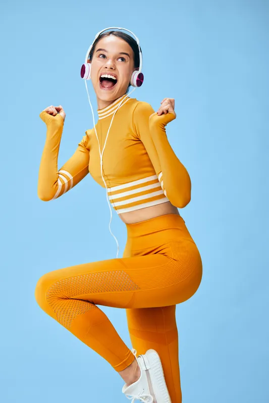 Fitness and sport themed portrait of a woman in bright orange activewear jumping with joy, wearing headphones, blue background, capturing energy, movement and motivation for training and wellness.