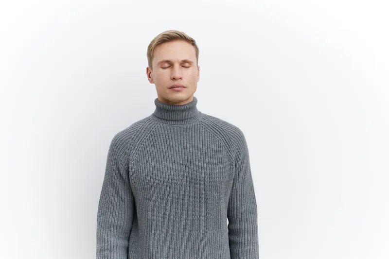 Close-up of a young man with short blonde hair and closed eyes, wearing a cozy gray knitted turtleneck sweater, standing against a plain white background, studio portrait, relaxed mood, fashion style.