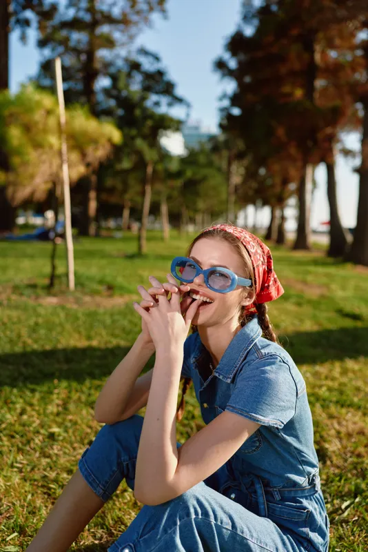 smiling woman wearing blue glasses and red headscarf sitting on green grass in sunny park with trees and relaxed atmosphere