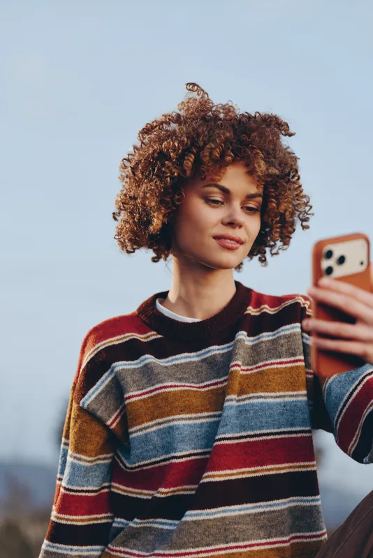 Woman selfie moment outdoors, curly hair framing her face, phone in hand, sweater portrait in casual lifestyle, soft daylight glow, relaxed smile, blue sky background, natural tones