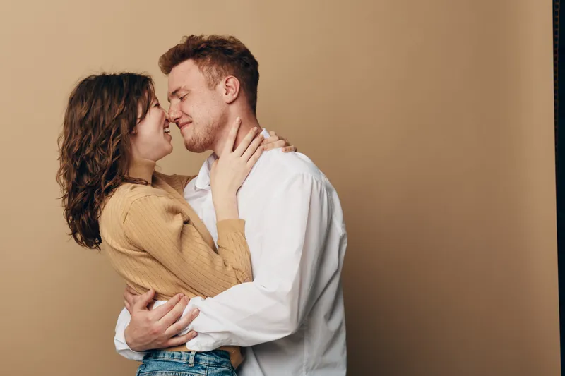 Young couple embraces closely with eyes closed, smiling gently in warm beige studio. Intimate moment of love, affection, and connection between man and woman in casual clothing.