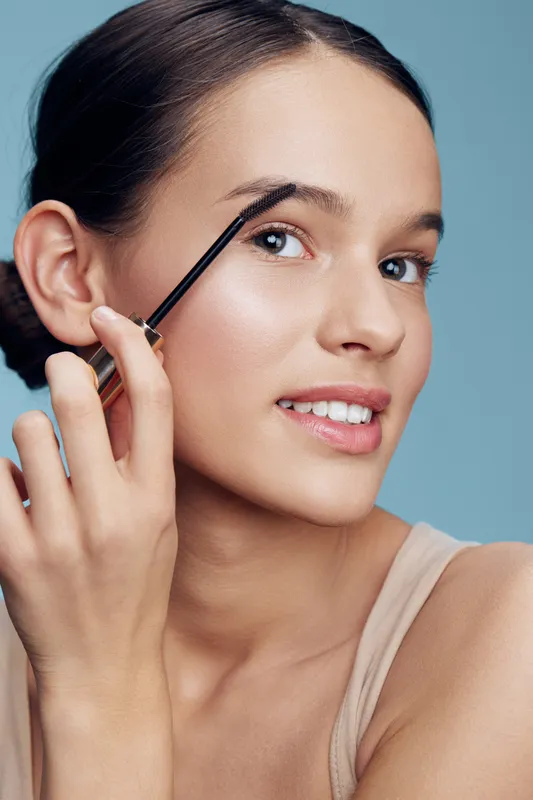 Young woman applying eyebrow gel, showcasing a natural beauty look with a soft smile, bright blue background, emphasizing personal grooming and self care