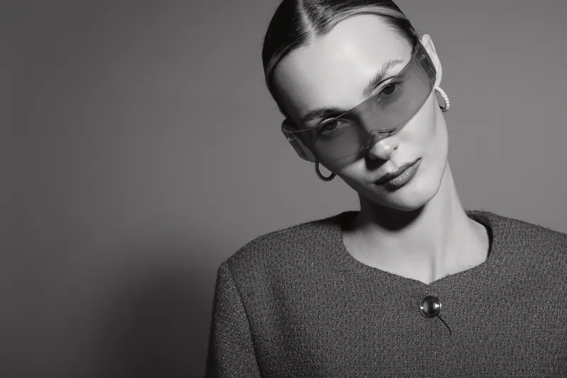 Elegant white woman wearing oversized sunglasses and hoop earrings poses confidently in a textured jacket against plain background. Concept of fashion and style.