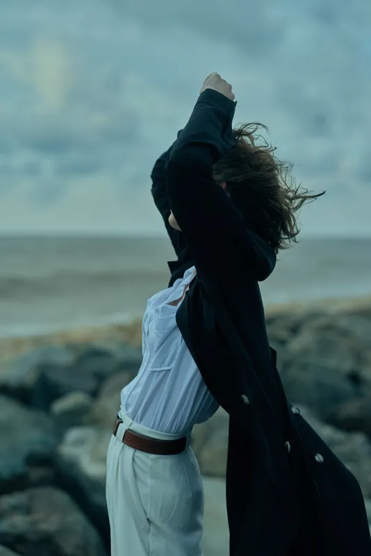 Windy outdoor moment by the sea with a figure lifting a coat over the head, casual white shirt and belt, rocky shoreline, gray sky, dynamic movement, mood of release and resilience
