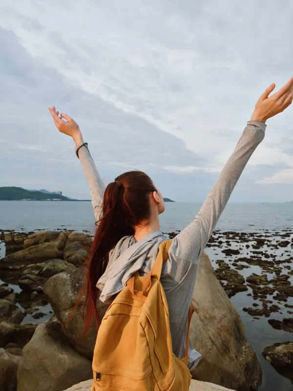 Young woman enjoying the ocean view with arms raised, wearing a casual outfit and a backpack, feeling free and connected to nature