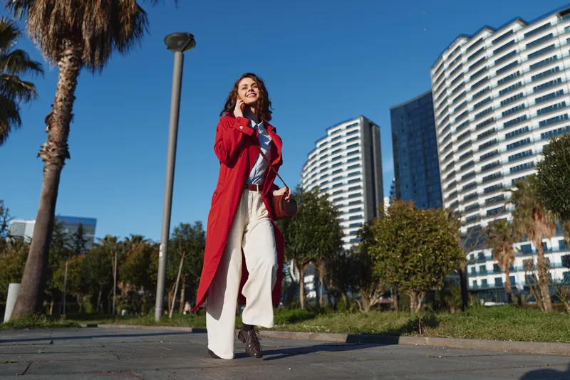 Young woman in red coat walks confidently on city street on sunny day with modern buildings and palm trees in background smiling happily.