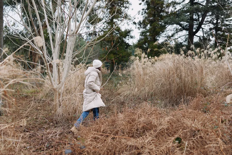 Person in a warm winter coat walks through tall dried grasses in a tranquil field, exploring nature and outdoor scenery with calm, muted tones and a quiet, reflective mood