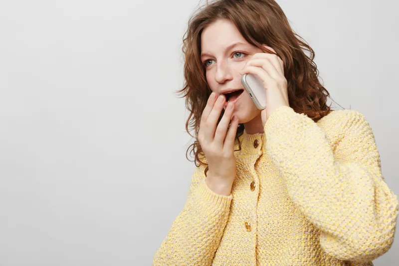 Surprised woman in yellow sweater talks on phone with open mouth and hand near face. Isolated on white background. Concept of unexpected news, communication, and emotional reaction.