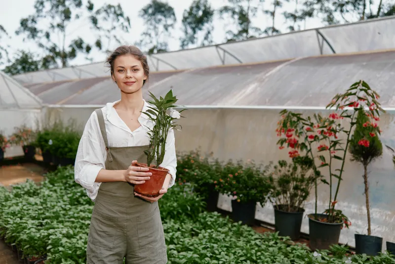 Young woman with a plant in a greenhouse, exuding joy and tranquility, dressed in casual overalls, surrounded by vibrant greenery and colorful flowers