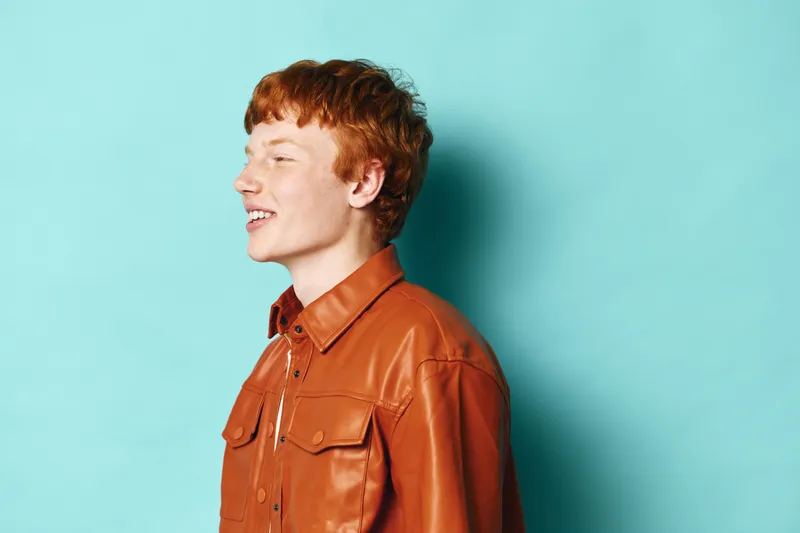 Young man with red hair wearing brown leather jacket stands against turquoise background smiling and looking sideways in casual style portrait photo with bright studio lighting.