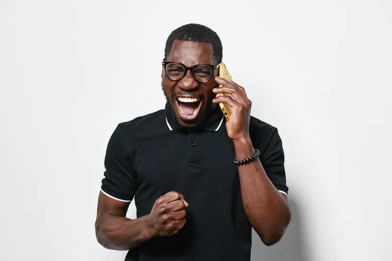 African American man with dark skin and glasses wearing black polo shirt talking on smartphone with excited expression and clenched fist isolated on white background in studio.