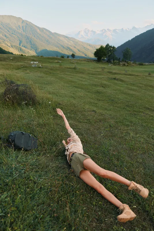 Young woman in a light beige dress lies on green grass in a wide mountain valley, enjoying a carefree moment as sunlight warms her relaxed posture and the peaceful surroundings