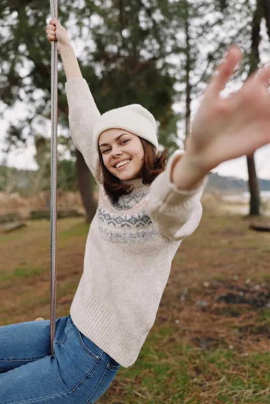 Woman on a swing outdoors in a cozy sweater and beanie, joyful and playful, smiling and reaching forward, surrounded by a calm autumn park setting.