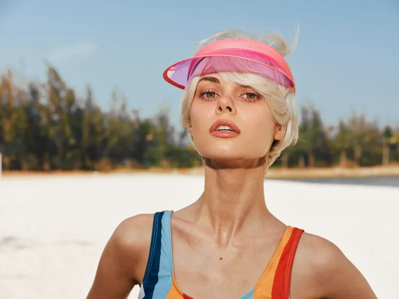 Portrait of a confident young woman wearing a translucent pink visor and a colorful swimsuit on a sunlit beach with clear sky and white sand, expressing vibrant energy.