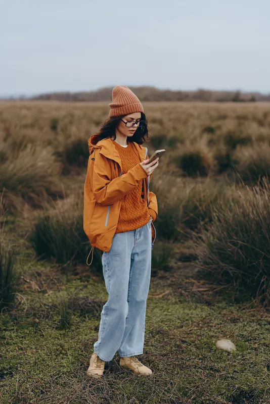 Outdoors lifestyle scene, young woman in orange jacket and knit beanie walk along a grassy path in a rural field, looking at a smartphone, calm mood, nature light, autumn