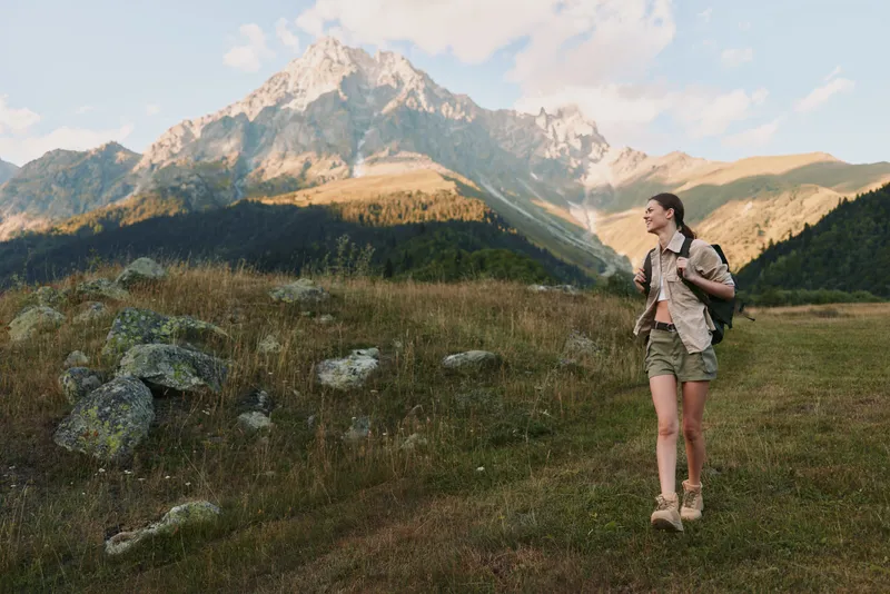 Young woman hiking outdoors in casual clothes with backpack walking on grassy trail near rocky mountain under clear sky, active lifestyle and nature exploration