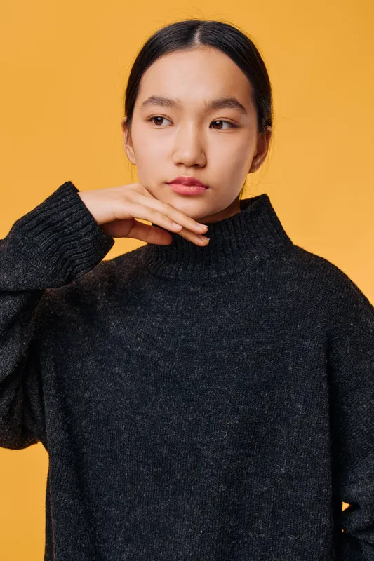 Young woman with dark hair in black sweater posing against orange background with thoughtful expression, hand near chin and looking sideways in studio portrait.