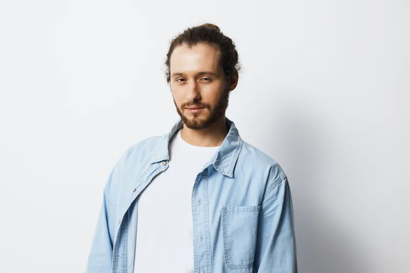 Young man with a casual look and confident expression in a denim shirt against a minimalist white background, ideal for lifestyle and fashion themes