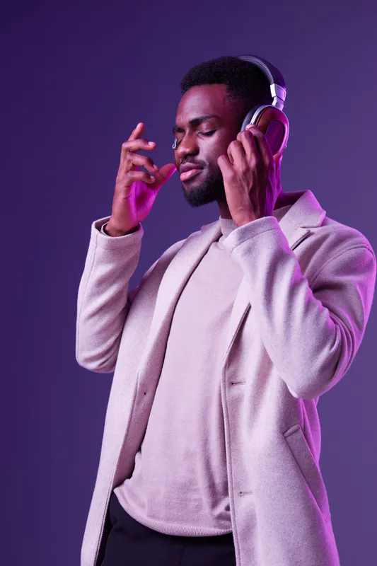 A stylish Black man enjoying music with headphones on a purple background, showcasing relaxation and confidence in modern fashion