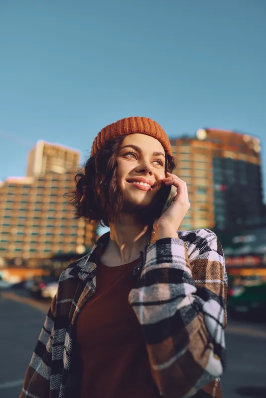 woman phone smile urban golden hour glow candid lifestyle and authenticity evoke mindful living; emotional storytelling portrait of a young woman in a beanie and plaid jacket outdoors