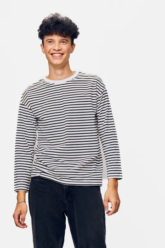 smiling man wearing striped long sleeve shirt and black pants stands against plain white background with cheerful expression and relaxed posture