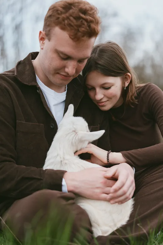 Smiling couple outdoors embracing a white goat kid, highlighting lifestyle, nature, and animal bonding. Soft natural light on a calm scene, creating a warm, affectionate mood. The scene emphasizes
