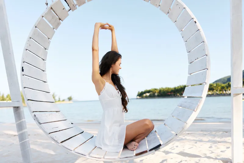 A serene woman in a white dress sits within a circular wooden swing by a sunlit beach, posing gracefully with arms raised, framed by blue sky and sea.