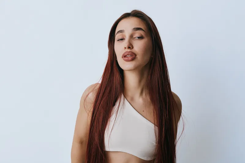 Young woman with long straight hair wearing white crop top posing against plain background, looking confident with closed eyes and relaxed expression in studio portrait.
