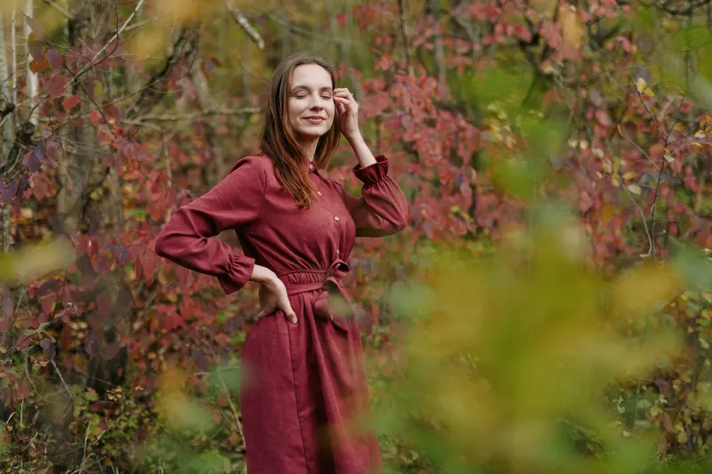 An authentic portrait of a woman in a burgundy dress stands among autumn foliage, conveying genuine confidence and natural beauty in a candid, grounded moment.