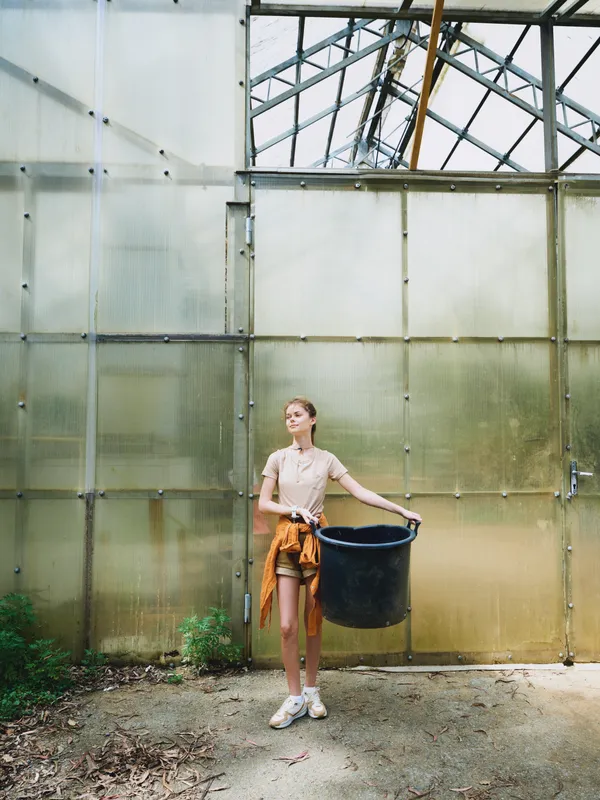 Woman greenhouse bucket gardening plants casual container garden seedling pot soil watering planter glass light sunlight young adult standing portrait jacket tied at waist indoor horticulture