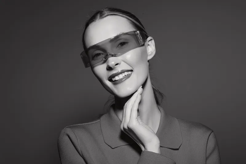 Smiling Caucasian woman with fair skin wearing futuristic glasses and a blazer touches her neck in a studio with dark background. Concept of fashion and innovation.