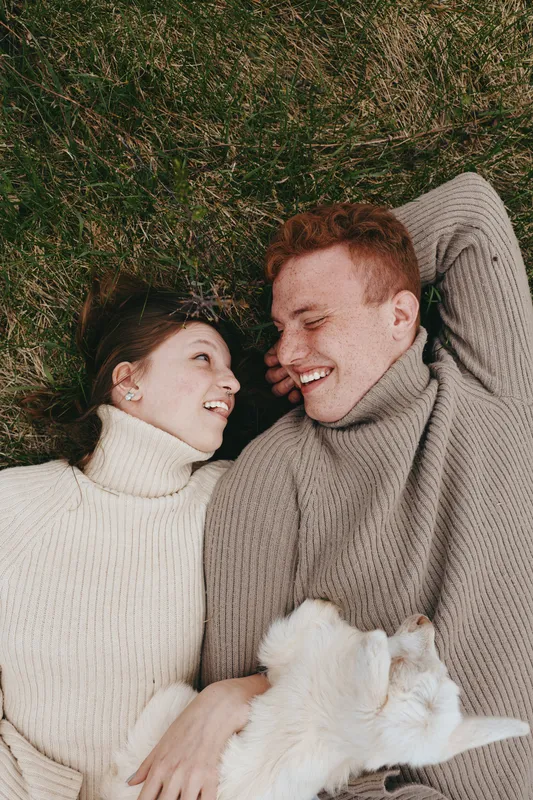 Smiling couple lying on grass in cozy sweaters with a goat kid nearby, enjoying a relaxed lifestyle moment outdoors. Natural light highlights warmth, connection, and wellness, creating a gentle sense