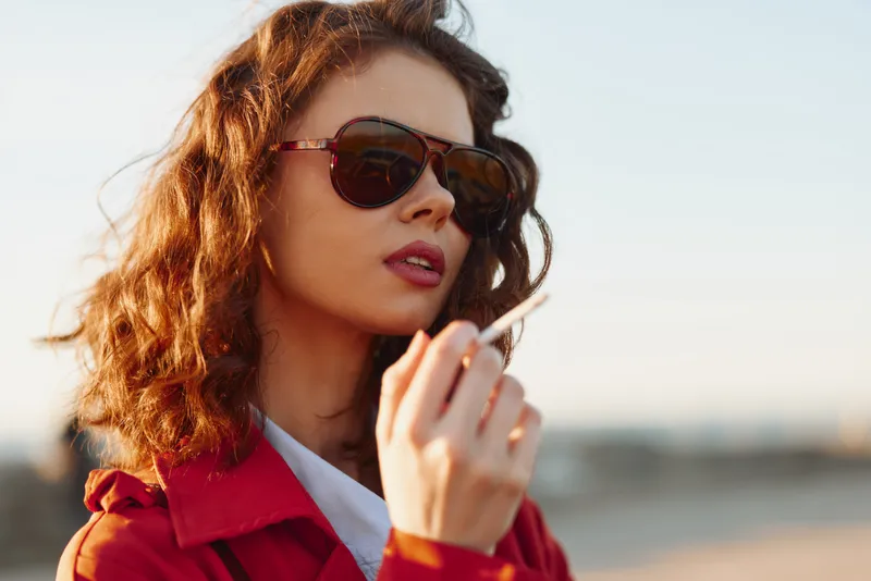 Young woman wearing sunglasses and red jacket outdoors, holding cigarette, natural light, curly hair, thoughtful expression, blurred background