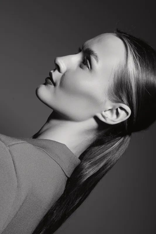 Profile of a young white woman with light skin and brown hair in a ponytail looking upward. Studio portrait with soft lighting and neutral background.