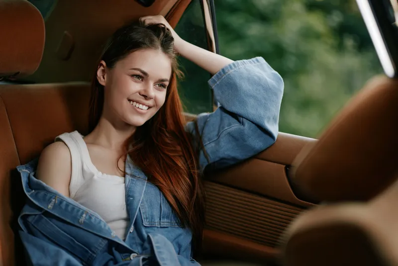 Young woman in a denim jacket smiling by the car window, enjoying the nature outside, showcasing a cheerful mood, summer vibe, and vibrant colors.