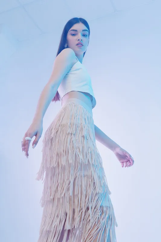 Young woman in a white crop top and fringed skirt poses confidently in soft blue studio lighting. Concept of woman beauty fashion with modern style and elegance.