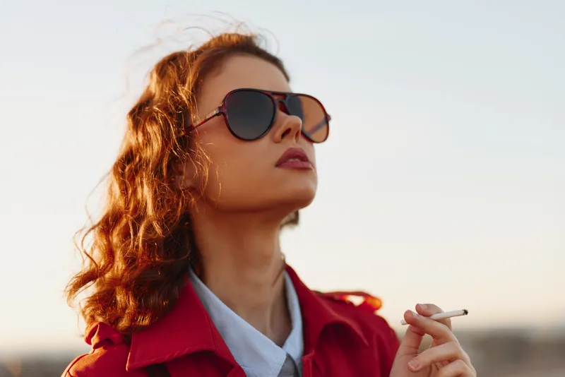 Stylish young woman with red hair wears sunglasses and a red coat outdoors in warm sunlight, smoking a cigarette and exuding confident relaxed mood as the breeze moves her hair.