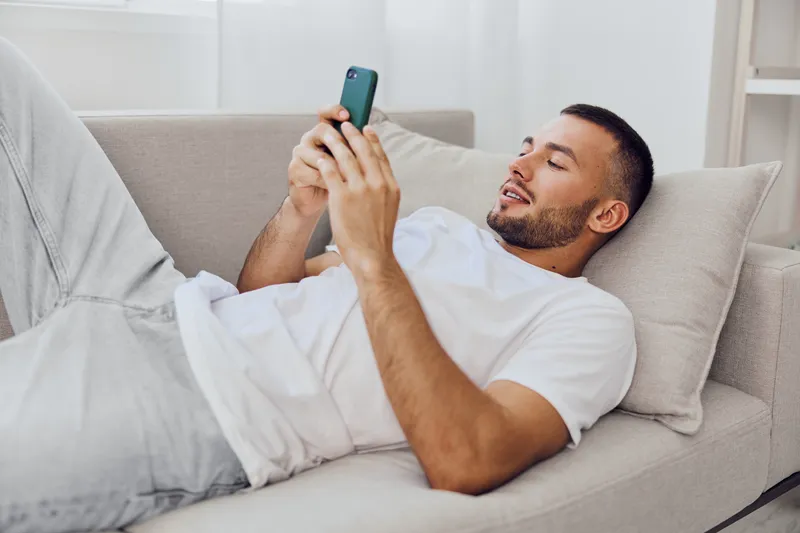 Relaxed man using smartphone on couch, enjoying leisure time with casual outfit and a cheerful mood, surrounded by a bright, modern interior.