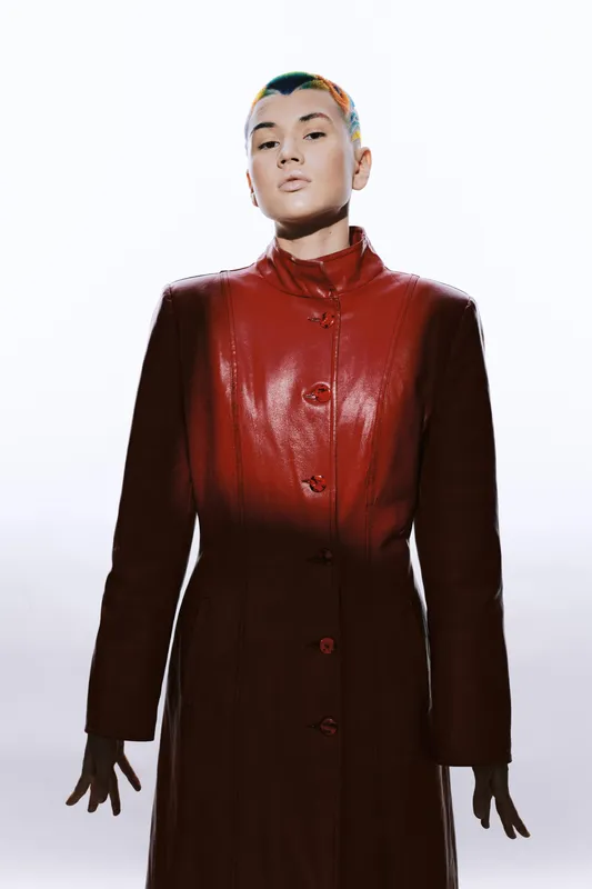 Young woman wearing a glossy red leather coat with a futuristic hairstyle, standing confidently against a bright white background in high fashion style.