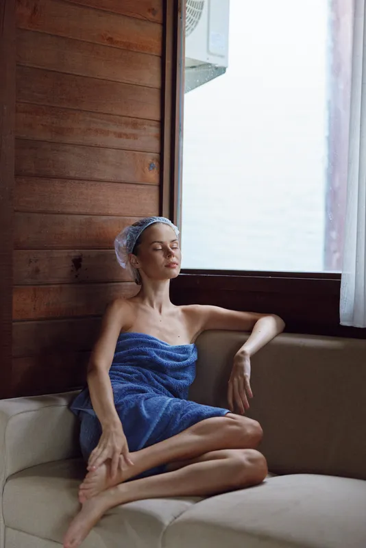 Relaxed woman wrapped in a blue towel sitting on a beige couch by a wooden wall and large window, enjoying calm and peaceful indoor atmosphere in soft natural light.